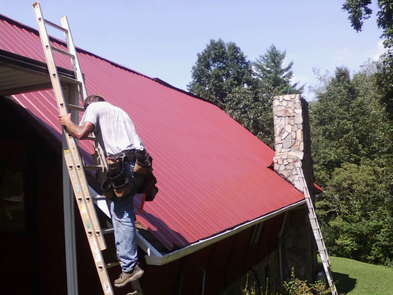 Roofer on a ladder beside a red metal roof and stone chimney — Soffit & Fascia Repair by Roofing Brothers in Thurman